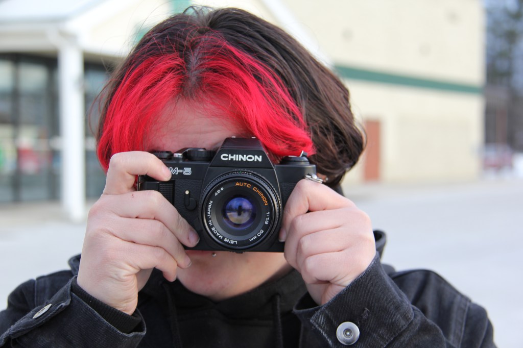 Image of a red haired person with a film camera covering their face.